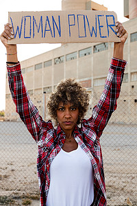 抗议者摄影照片_自信的非裔美国人示威抗议者看着相机，头顶举着一个女性权力标志。