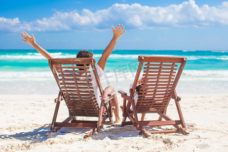 带着小女儿坐在沙滩椅上的年轻父亲在岸边的海洋上举起双手