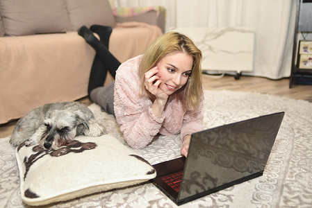 穿着粉色毛衣的可爱女人躺在地毯上，家里有一台笔记本电脑，一只灰色的狗睡在她旁边的枕头上。
