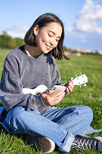读后感背景图摄影照片_用智能手机微笑的亚洲女孩的垂直照片，弹奏四弦琴，边唱歌边读和弦或歌词，在户外放松。