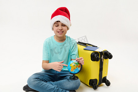 一个戴着圣诞帽的小男孩的孤立肖像，指着地球上的旅游目的地，坐在黄色手提箱旁边。