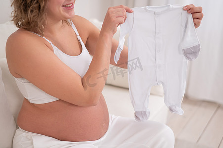 待产摄影照片_一位孕妇坐在白色沙发上，拿着待产婴儿的衣服。