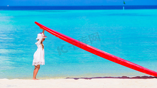 白色冲浪板摄影照片_热带白色沙滩上带红色大冲浪板的可爱小女孩