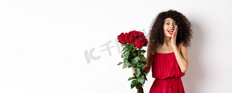 身着红色裙子、兴奋的卷发女人，收到一束玫瑰，看起来很惊讶，为浪漫的礼物而欢欣鼓舞，站在白色背景上