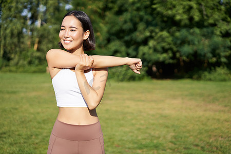 亚洲女性伸展双臂，在公园里做健身运动，开心地笑着，热身，然后在白天呼吸新鲜空气慢跑