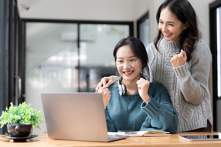 两名年轻的亚洲女性在现代办公室里用笔记本电脑开心地微笑着，表现出工作成功的喜悦表情。