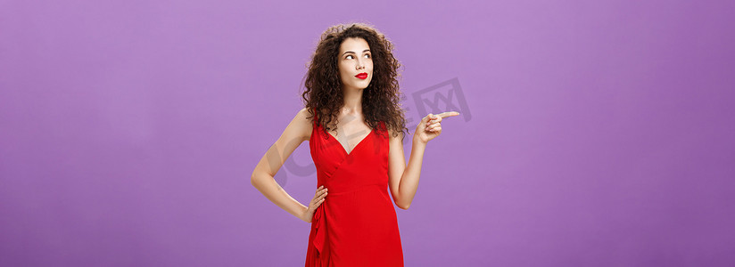 身着优雅红色晚礼服、留着卷发的体贴而梦幻的迷人女性的画像，她指着左手，好奇地握着腰，在紫色的墙上摆出女性化的姿势