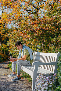 公园里的长椅摄影照片_一个少年坐在秋天公园的长椅上，喝着保温杯里的咖啡，看着手机。