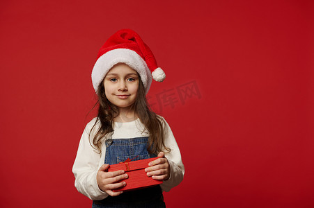 拆圣诞节礼物摄影照片_可爱的小女孩戴着圣诞老人的帽子，带着可爱的礼品盒，在红色背景上为圣诞节带来惊喜。