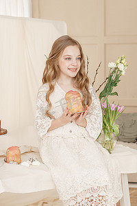 一个长头发、穿着浅色连衣裙的女孩坐在复活节餐桌旁，桌上放着蛋糕、春花和鹌鹑蛋。