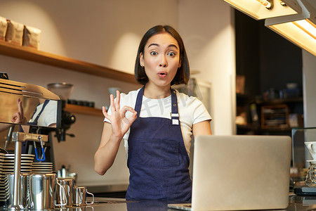 咖啡师女孩的画像，亚洲女咖啡馆工作人员，表现得很好，可以签署批准，推荐 smth，快乐和高兴，穿着围裙，站在柜台后面拿着笔记本电脑