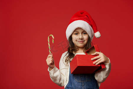 快乐的小女孩对着镜头微笑，拿着糖果和礼盒，里面装着快乐的圣诞礼物，与世隔绝在红色背景中