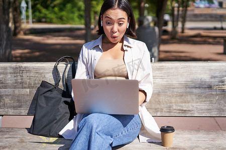快乐的亚洲女孩聊天，在笔记本电脑上进行视频通话，坐在公园的长椅上交流，与网上的人交谈