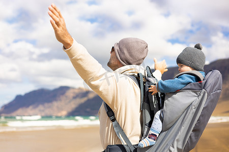 在西班牙兰萨罗特岛 Famara 多风的沙滩上，年轻的父亲在背包里背着他的婴儿男婴太阳享受纯净的大自然，同时举起双手向天空。