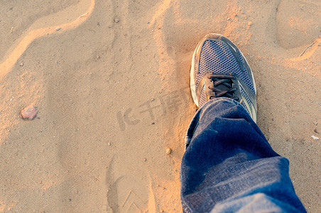运动鞋展示摄影照片_从上往下看男人用一条破旧的运动鞋牛仔裤在尘土飞扬的沙滩上伸出一只脚，展示了晚上旅行和徒步旅行，因为阳光从它身上反弹