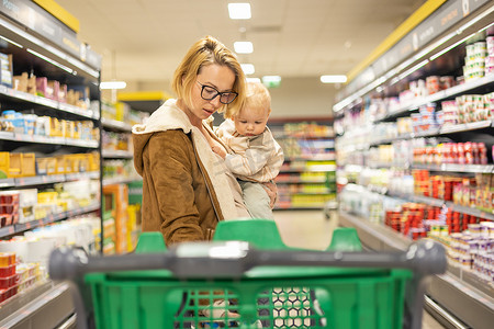 母亲带着婴儿男婴购物，将购物车推到超市杂货店的部门过道。