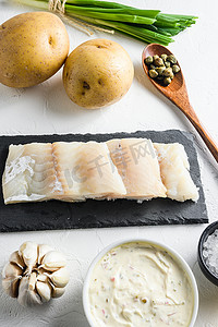 传统的鱼和薯条配料配方石板面糊上的生鳕鱼片、土豆、塔塔酱、柠檬、酸豆、绿色香草大蒜、盐、白石背景侧视图上的胡椒粒垂直特写