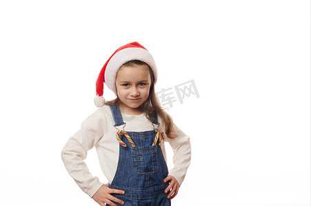 圣诞白色背景摄影照片_可爱的小女孩戴着圣诞帽，在他的牛仔工作服的口袋里摆着糖果手杖，白色背景