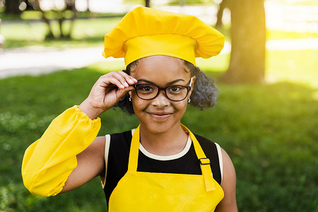 穿着厨师帽和黄色围裙制服的非洲黑人儿童厨师女孩摸着 hil 眼镜，在户外微笑。