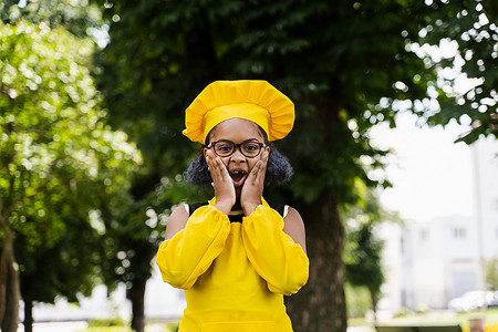 身穿厨师帽和黄色围裙制服的黑人非洲儿童厨师女孩震惊地双颊和惊喜。