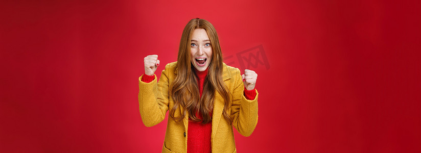 冬天红墙摄影照片_Studio shto 身穿黄色外套的快乐兴奋的漂亮姜女郎在快乐和幸福中举起紧握的拳头，从成功和胜利中大喊是，庆祝战胜红墙