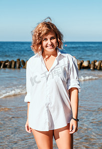 海边女子摄影照片_身穿白色泳衣和衬衫的年轻金发女子站着