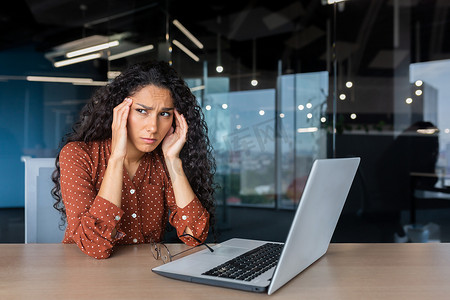 西班牙女摄影照片_在工作中过度疲劳的西班牙裔女性，戴着眼镜的女商人手举着头，头痛严重，在办公室里工作，坐在桌旁，在工作时使用笔记本电脑