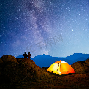 年轻夫妇坐在灯火通明的帐篷附近的岩石上，在美丽的山脉上方观看明亮的银河