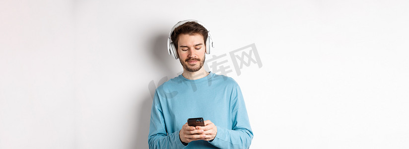 身穿蓝色运动衫的现代年轻人在智能手机上放音乐，用无线耳机听歌，白色背景