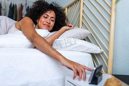 睡觉觉摄影照片_非洲裔美国年轻女性在睡了一个好觉后醒来时很开心，她伸出手来关掉闹钟。