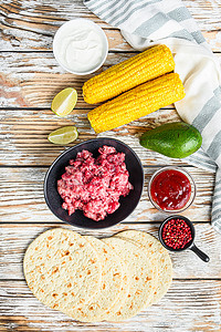 用黑色碗中的碎有机牛肉、玉米、卡尔萨在白色纹理木桌上烹制墨西哥炸玉米饼配料，俯视图。