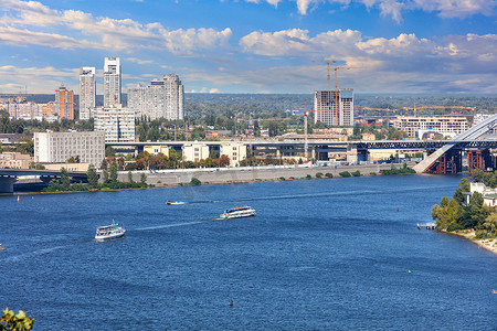游船河摄影照片_在明亮、美丽的天空和城市建筑的背景下，游船沿着基辅的城市堤岸沿着第聂伯河的水面行驶。