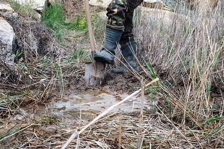 挖矿山摄影照片_穿着橡胶靴的男人在沼泽地里挖了一个铲坑。