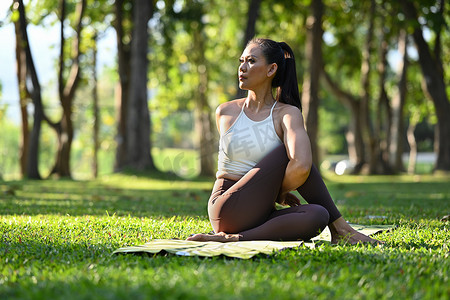 阳光下的女性摄影照片_穿着运动服的优雅千禧一代女性坐在半鱼王的姿势中，在阳光下的绿色公园里练习瑜伽