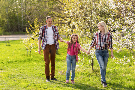 户外肖像快乐的年轻家庭在盛开的树下的春天公园玩耍，可爱的夫妇和小孩在阳光明媚的花园里玩得开心