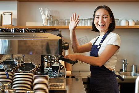 热情的亚洲咖啡师，女孩用咖啡机，向客户挥手，打招呼，在她的咖啡馆里问候客人