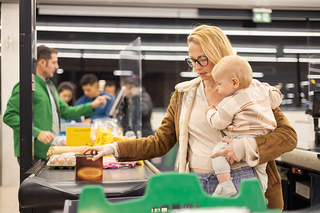 母亲带着婴儿男婴购物，抱着孩子在超市杂货店的收银台堆放产品。