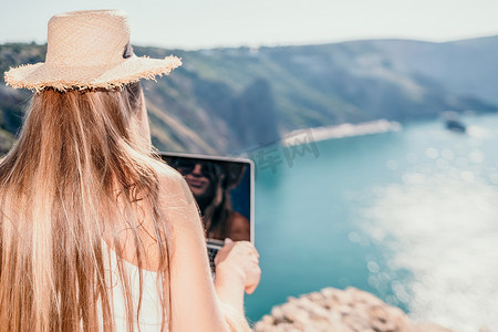 坐在海边摄影照片_数字游牧者，戴帽子的女人，日落时分坐在海边岩石上的带笔记本电脑的女商人，从远处在网上进行商业交易。