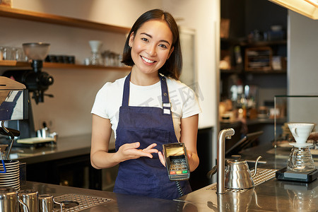 亚洲咖啡师女孩在柜台的画像，向想要非接触式支付、点菜、站在咖啡馆的客户展示卡片机