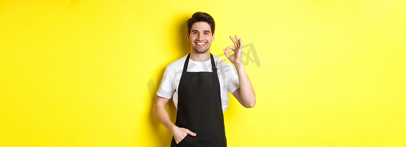 身着黑色围裙、微笑的年轻咖啡师站在黄色背景上，展示着好的标志，推荐咖啡店或餐厅