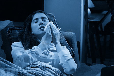 坐在沙发上的年轻女性，身上盖着一条毯子，冻得鼻涕直流，发烧、被抓、生病的女孩有流感症状、流感或病毒概念
