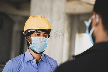 现场的两名建筑工人或工程师戴着医用面罩交谈，同时保持社交距离 — 商业概念、行业重新开放或工作场所的 covid-19 安全措施。