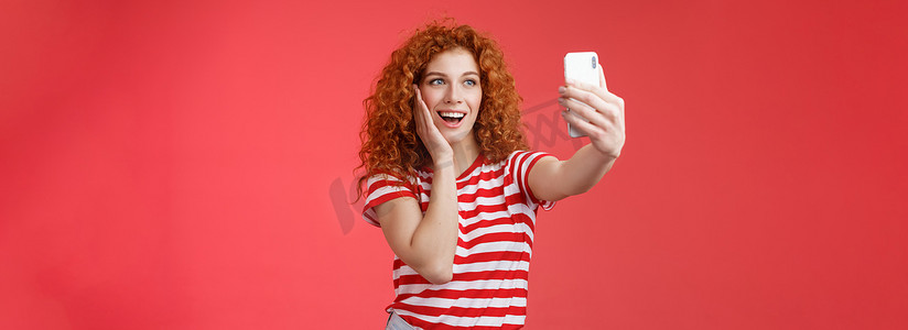 时尚可爱的红发卷发女性夏季 T 恤录制视频社交媒体，来自豪华热带度假村出国旅游度假自拍手持智能手机摆出愚蠢的照片红色背景