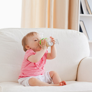 可爱的金发婴儿坐在沙发上用奶瓶喂养