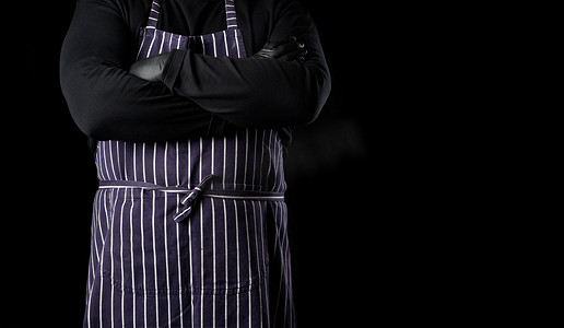 身穿蓝色条纹围裙和黑色衣服的男厨师站在黑色背景下