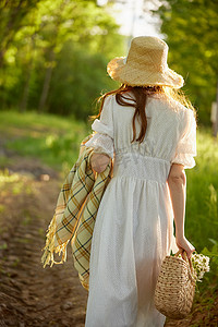 在阳光明媚的天气里，一个身穿浅色连衣裙、头戴柳条帽、手拿篮子和格子花呢的女人穿过森林。