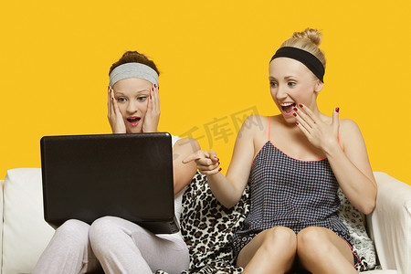 黄色背景下坐在沙发上使用笔记本电脑的两名震惊的年轻女性