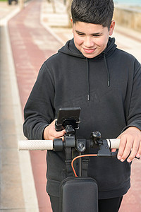 14-17 岁的前视图在自行车道上快乐地微笑着骑电动滑板车，并使用智能手机音乐应用程序，E-Mobility 城市休闲概念