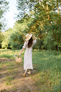 一位身穿夏装长裙的女士在林间小径上行走，手上戴着一顶柳条帽
