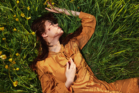 红色长围裙摄影照片_一个长着红色长发的平静女人躺在黄色花朵的绿色田野里，穿着橙色的连衣裙，闭着眼睛，把手放在脸旁，享受着平静和休养